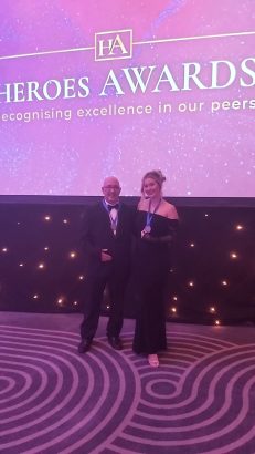 Two people in black tire attire stand in front of a backdrop that reads Heroes Awards