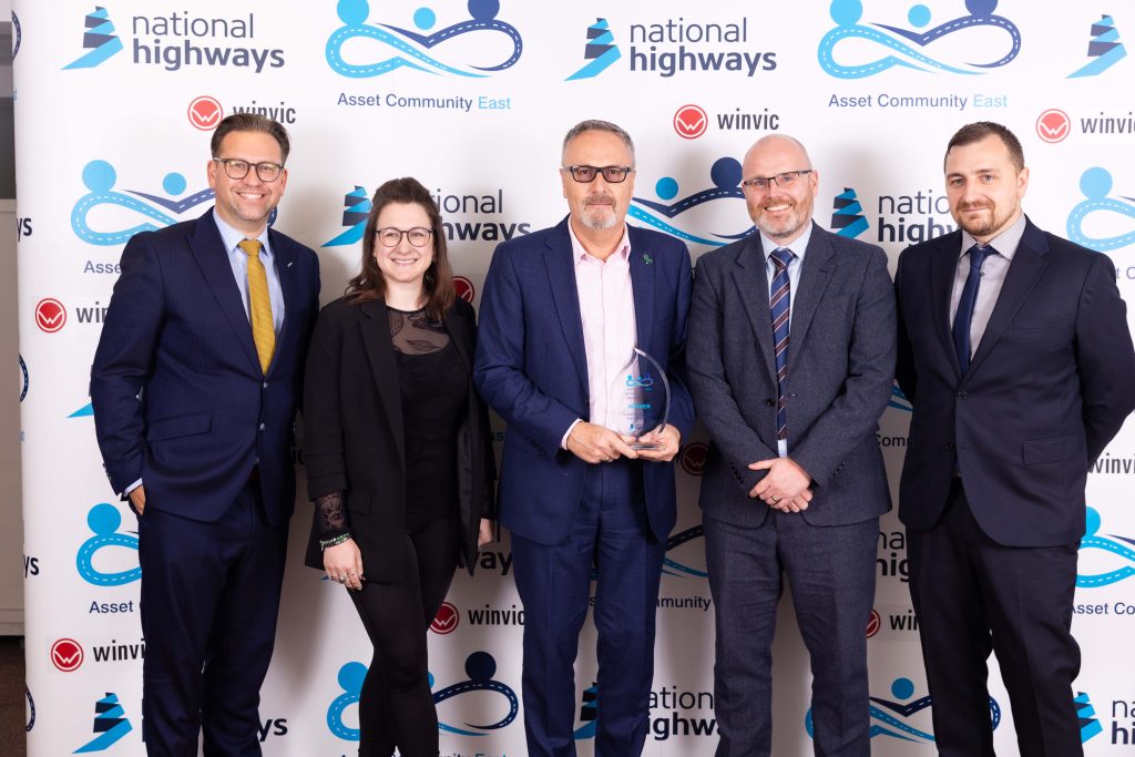 A group of 5 people stand in front of a background featuring the National Highways and ACE Community Logos. Second from the left is HW Martin’s Group Marketing and Communications Manager Joanna Hill who presented the award to the winners Ringway Infrastructure Services