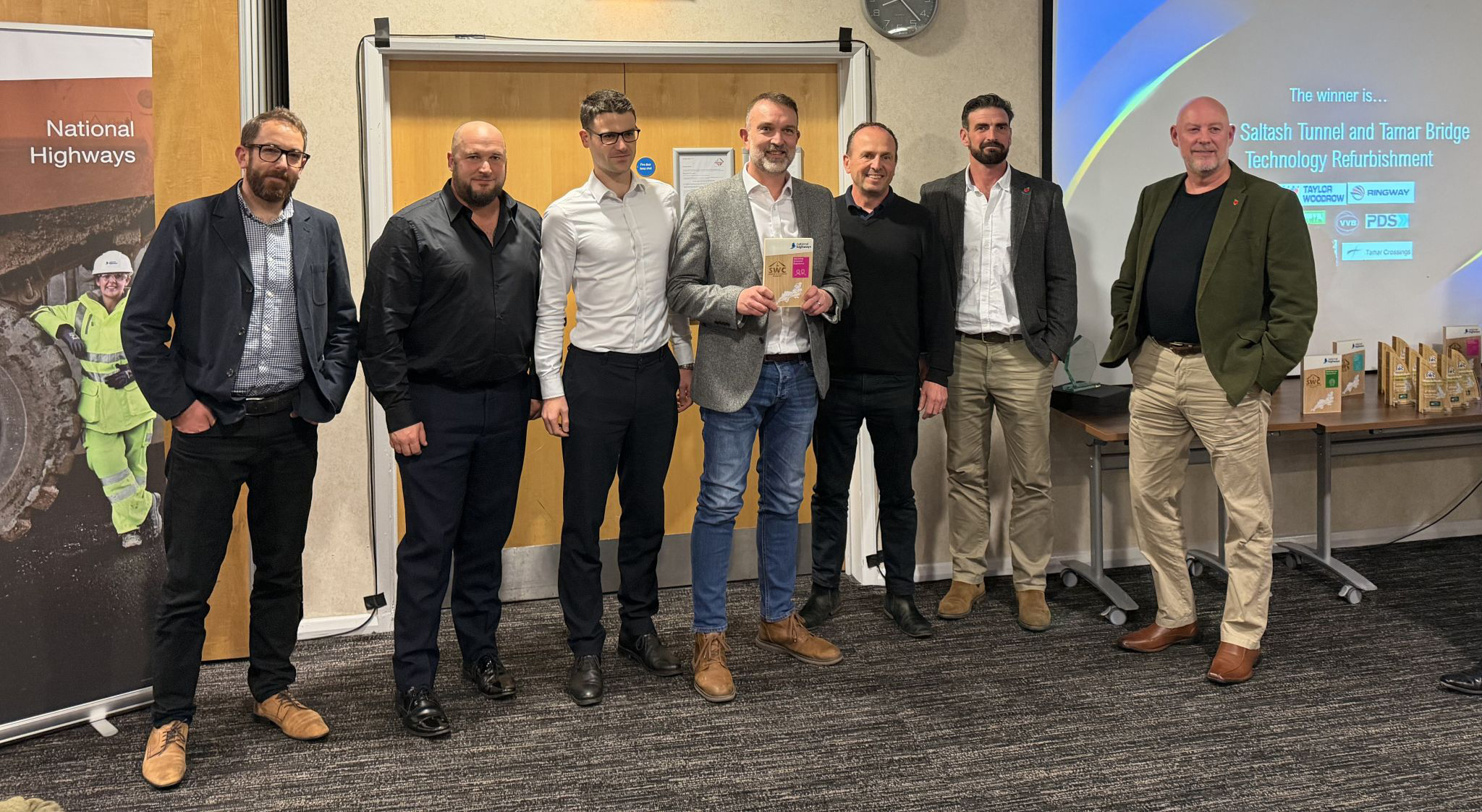The image shows seven individuals standing side by side in a conference or meeting room. They are dressed in business or smart-casual attire, including blazers, shirts, trousers, and shoes. One person in the center is holding an award or plaque. In the background, there is a wooden door with a light-colored frame. To the left of the group, there is a vertical banner displaying the National Highways logo and an image of a person wearing high-visibility safety gear. On the right side of the image, a large presentation screen is visible. The screen displays text that reads: “The winner is… Saltash Tunnel and Tamar Bridge Technology Refurbishment” Several logos are also visible on the screen, including National Highways and others related to the project. In front of the screen, there is a table with multiple brown paper bags and small boxes arranged neatly, suggesting event giveaways or promotional materials. The floor is carpeted in a dark patterned design, and the walls are light-colored, creating a professional setting.