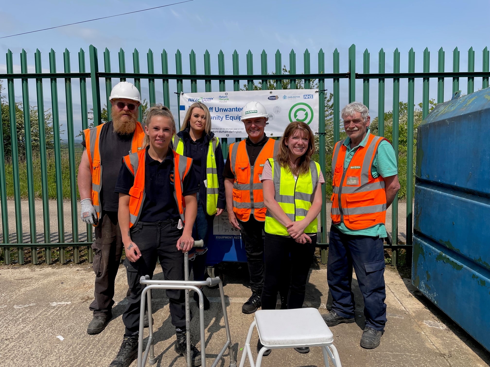 A photo features six people standing outdoors in front of a green metal fence and a banner that reads “North East Reuse Project – Medequip.” They are wearing high-visibility vests and work uniforms, and mobility aids such as a walking frame and a shower stool are placed in front of them.