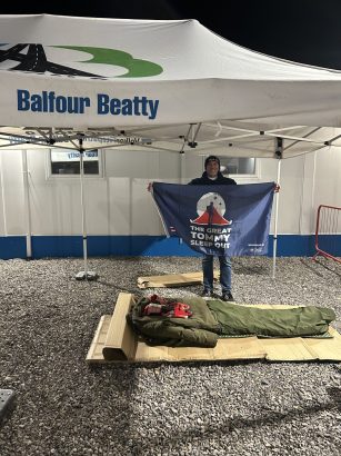 A man stands under a Balfour Beatty-branded canopy at night, holding a navy banner that reads ‘The Great Tommy Sleep Out.’ In front of him on the gravel ground is a cardboard base with a green sleeping bag and clothing laid out.