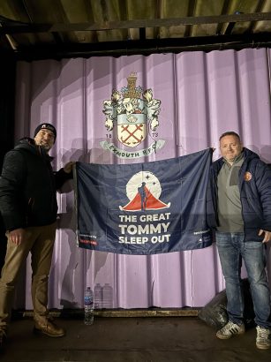“Two men stand outdoors at night holding a large navy banner that reads ‘The Great Tommy Sleep Out.’ They are positioned in front of a purple wall displaying the Exmouth RFC crest. A water bottle sits on the ground below the banner.”
