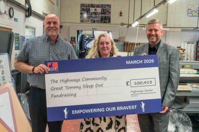 Left to right: Andy Graham, HW Martin Operations Director, Donna Dind and Andrew Stephenson from National Highways pose with an oversized RBVE cheque during a cheque presentation at a workshop facility. The cheque reads "The Highways Community Great Tommy Sleep Out Fundraising" and shows a total of £100,413 raised by the highways sector. The cheque is dated March 2026 and features the RBVE logo and the phrase "Empowering Our Bravest". Industrial machinery, workbenches and production equipment are visible in the background.
