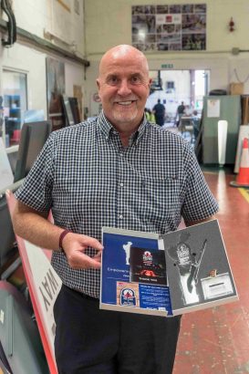 Andy Graham, Operations Director at HW Martin (Traffic Management) stands in an industrial workshop holding an RBVE commemorative presentation box. The open display contains Great Tommy Sleep Out memorabilia, including a thank you card, badge, engraved Tommy silhouette award and keepsake items recognising support for the veterans' fundraising initiative. Workshop equipment and signage are visible in the background.